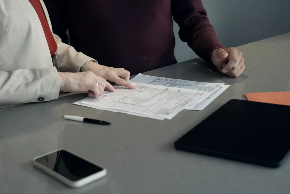 Two people at a grey table. One points to a line on a printed form while the other looks on.