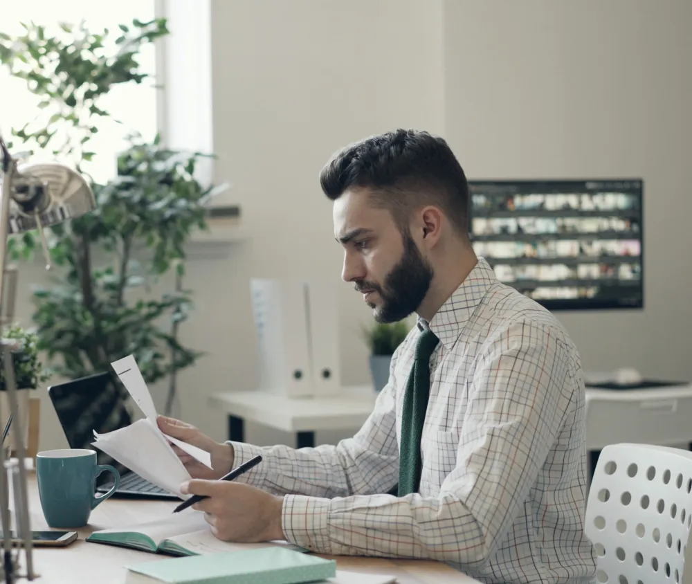 A man in a shirt and tie reads a printed document at his desk, with a laptop open beside him.